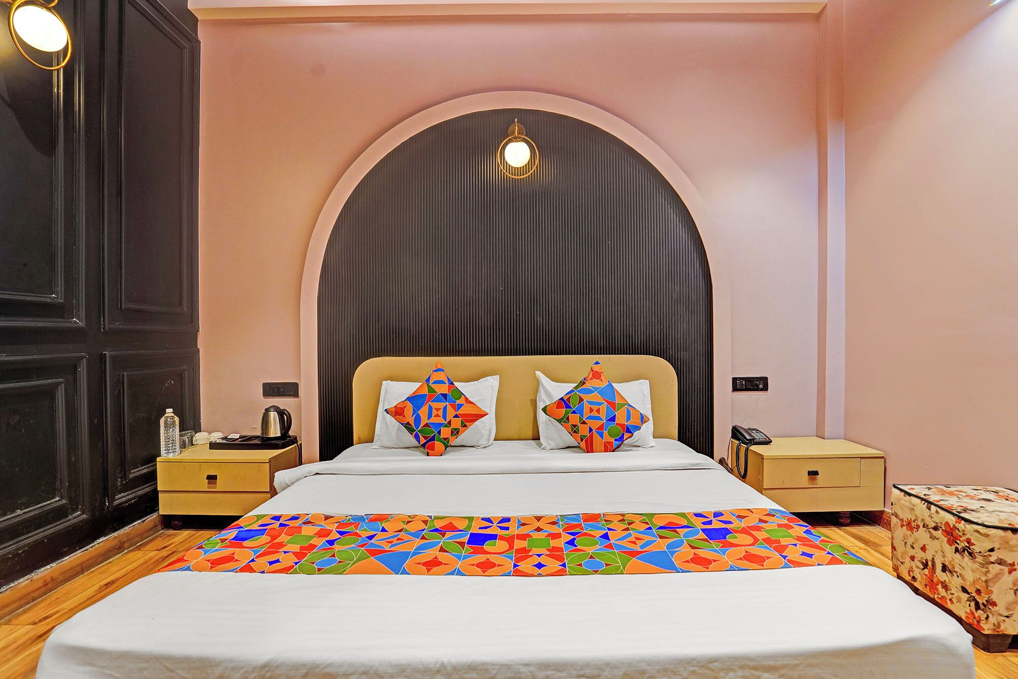 A hotel room with a bed, two bedside tables, a nightstand, a decorative rug, and a wall-mounted light fixture.