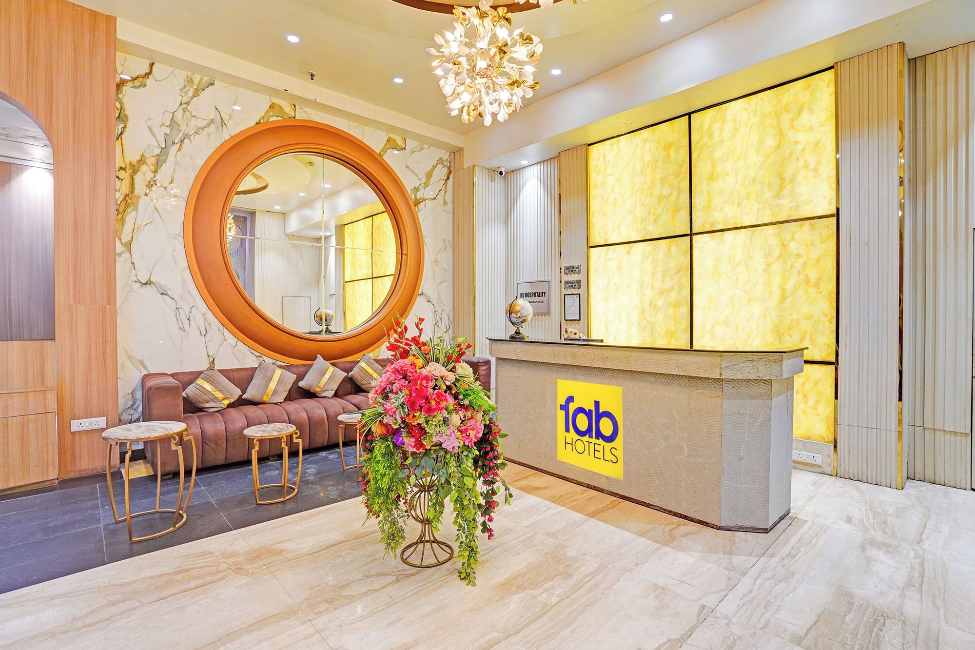 A hotel reception area with a round mirror, a sofa, a desk with a 'fab hotels' sign, and a large floral arrangement.