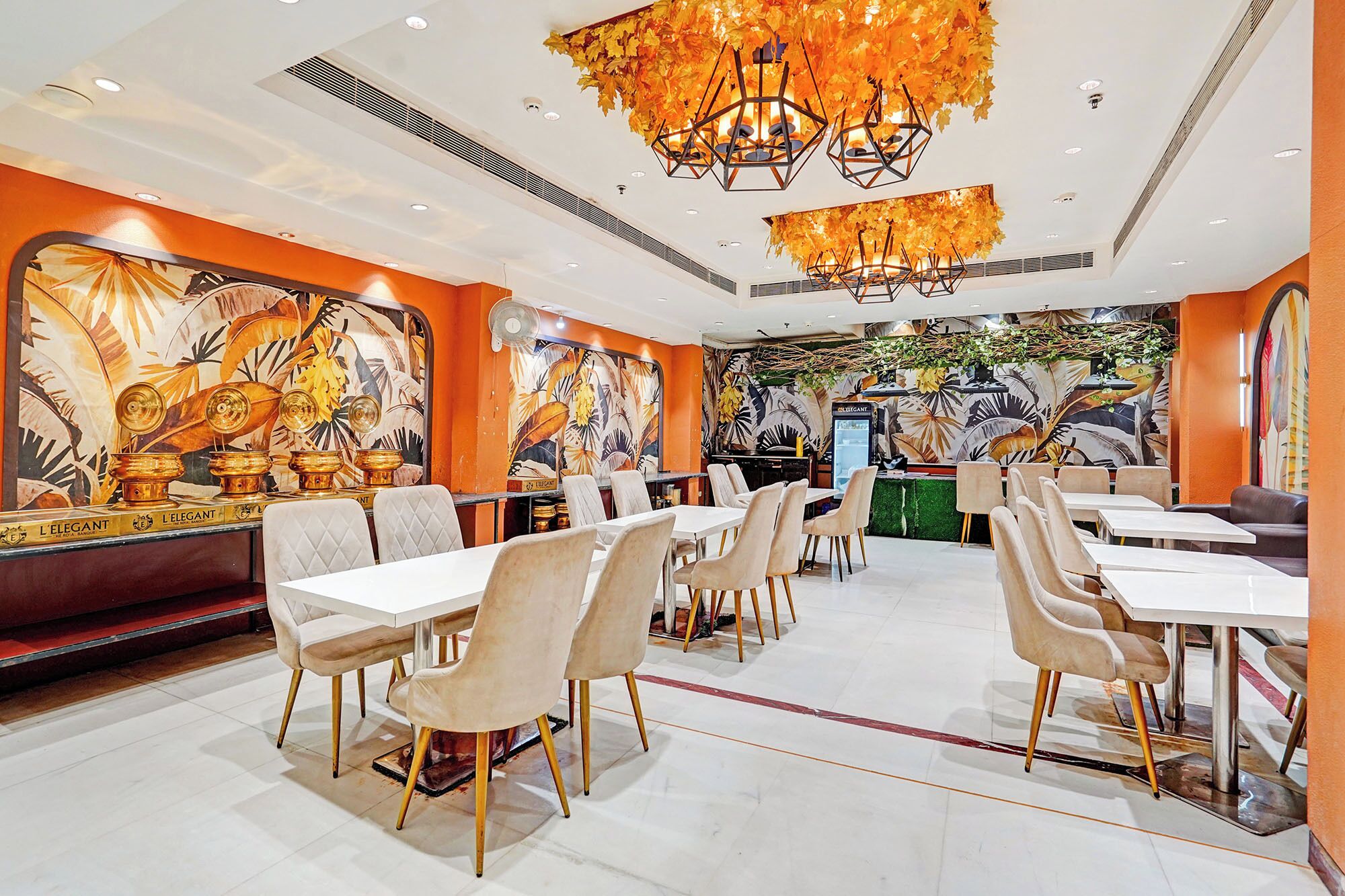 Un restaurante con una decoración moderna, que cuenta con un área de comedor central con mesas y sillas blancas, una zona de bar y un mural con motivos tropicales en las paredes.
