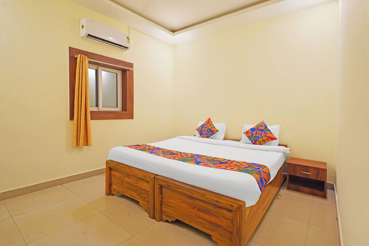 A neatly made bed with a patterned bedspread, a wooden headboard, a nightstand with a drawer, a wall-mounted air conditioner, and a window with curtains.