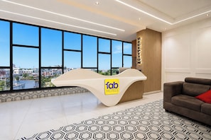 A hotel reception desk with a logo, a large window with a city view, a patterned rug, and a sofa with a red pillow. - FabHotel A Square - 400m from Bhimavaram Junction (Bhimavaram)