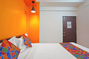 Habitación de hotel con pared naranja, cama con ropa blanca, manta de diseño geométrico y colores vivos, puerta oscura con un cartel, y lámpara colgante.