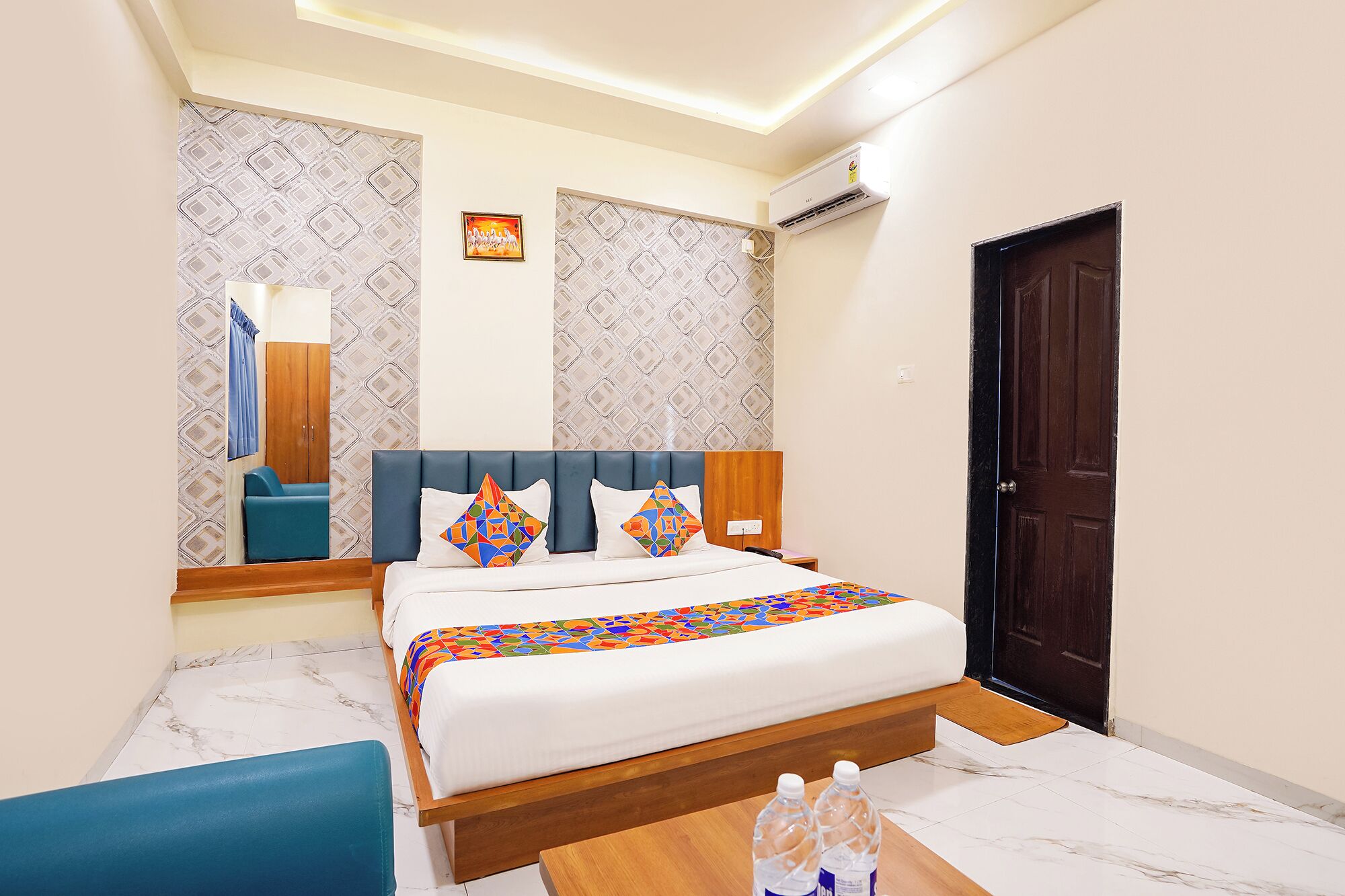 A hotel room with a bed, a wooden nightstand, a blue chair, a wooden door, and patterned wallpaper.