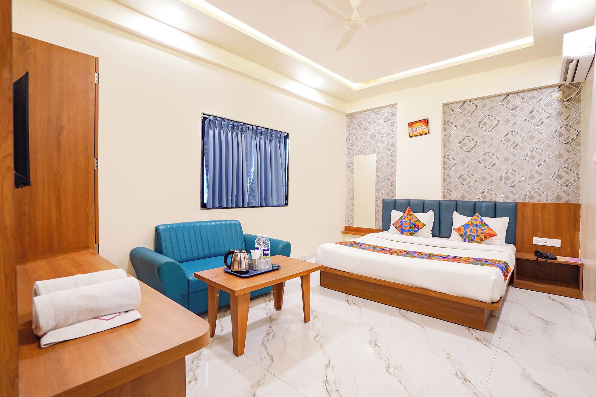 A hotel room with a bed, a blue sofa, a wooden table, a small table with a kettle, and a ceiling fan.