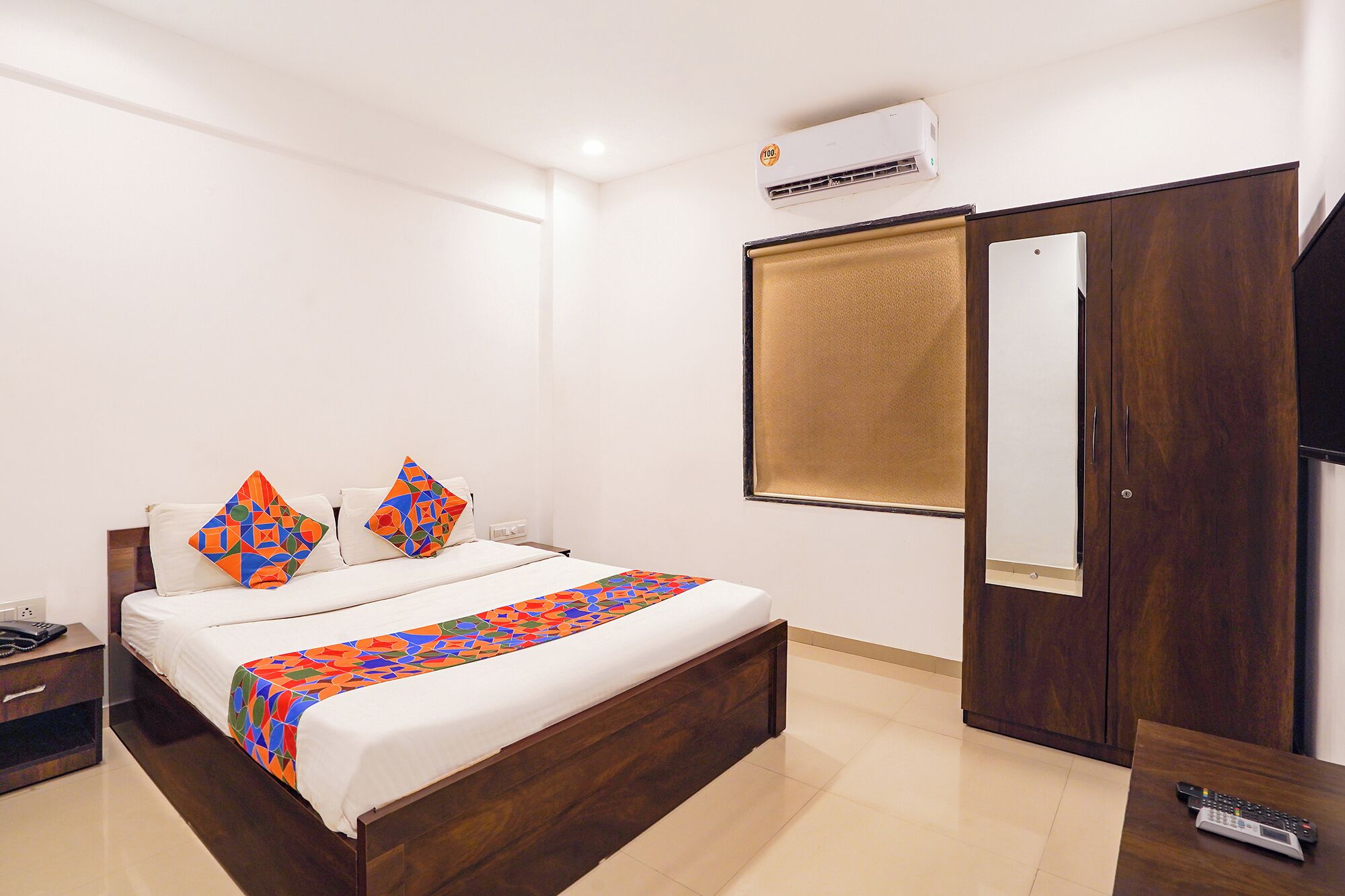 A hotel room with a bed, a bedside table, a wardrobe, and an air conditioning unit.