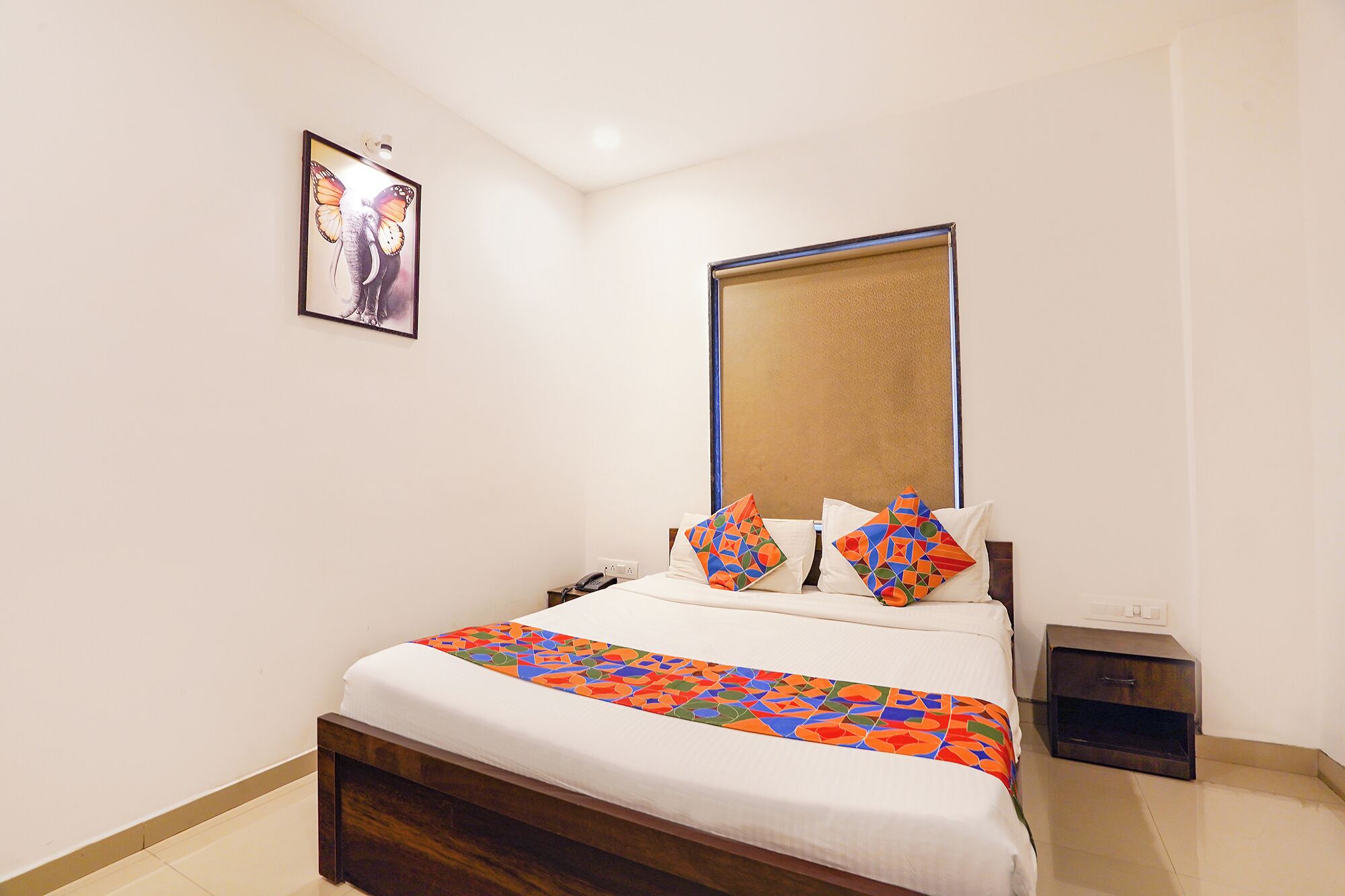 Una habitación de hotel con cama, mesita de noche y un espejo montado en la pared, además de un cuadro enmarcado.