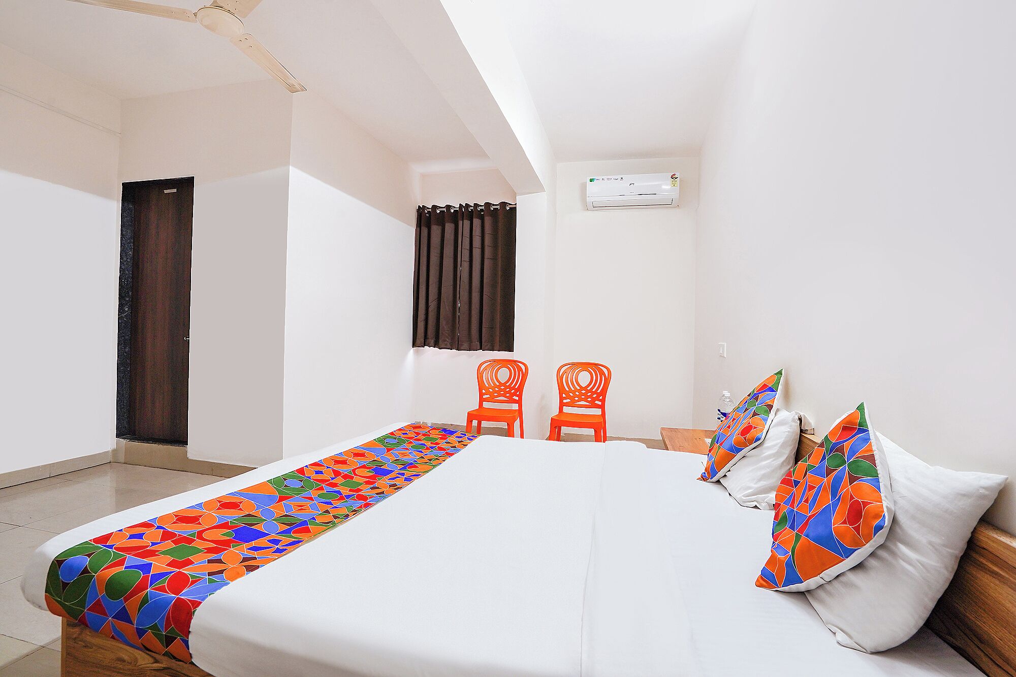 Une chambre d’hôtel avec un lit, deux fauteuils orange, une fenêtre avec des rideaux et un appareil de climatisation.