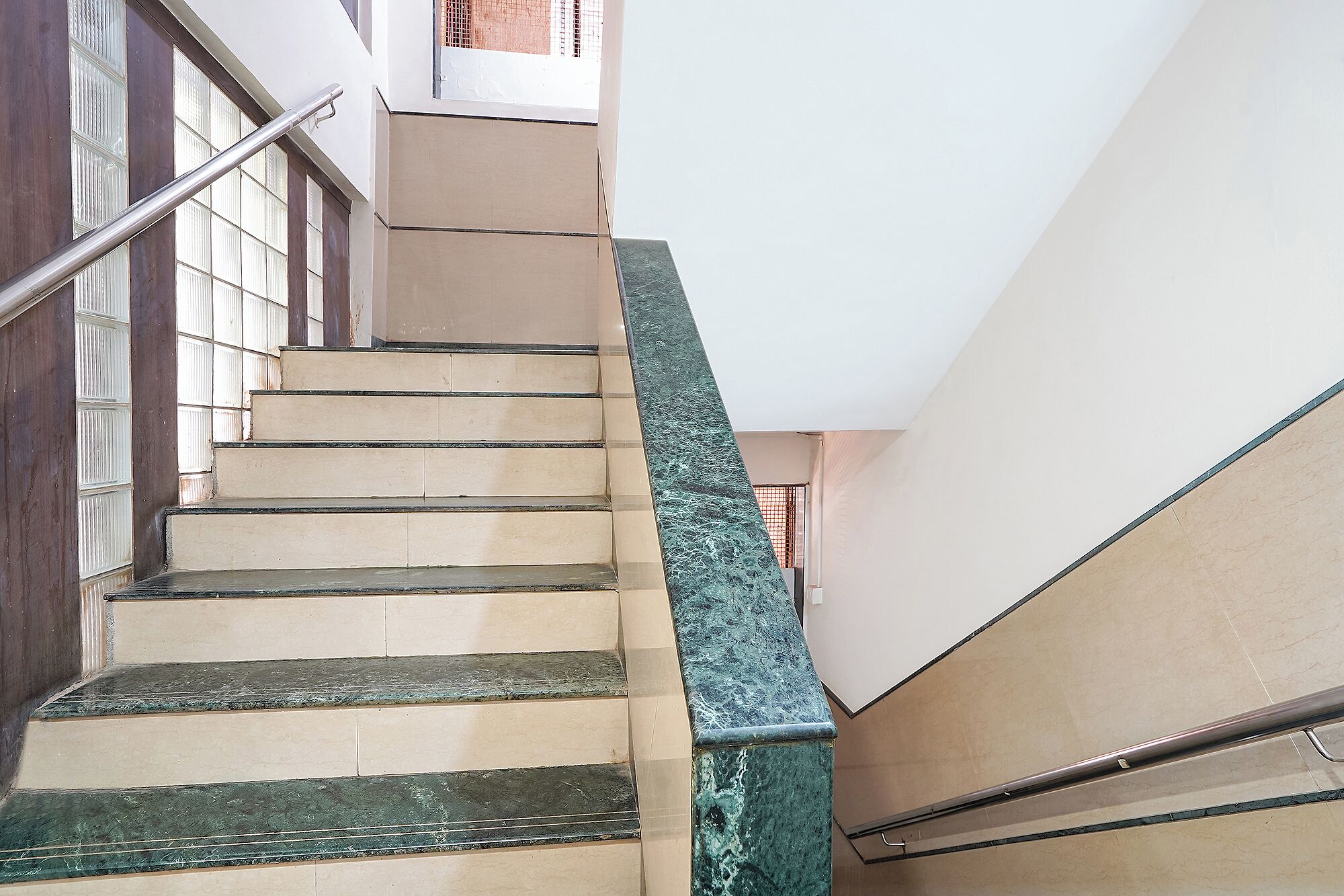 Eine Treppe mit beigen Stufen und einem grün marmorierten Geländer, die zu einem Zwischenpodest mit weißer Wand und einem Fenster führt.