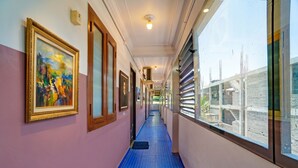 Un pasillo con obras enmarcadas en las paredes, piso de azulejos azules y ventanas amplias que ofrecen vistas al exterior.