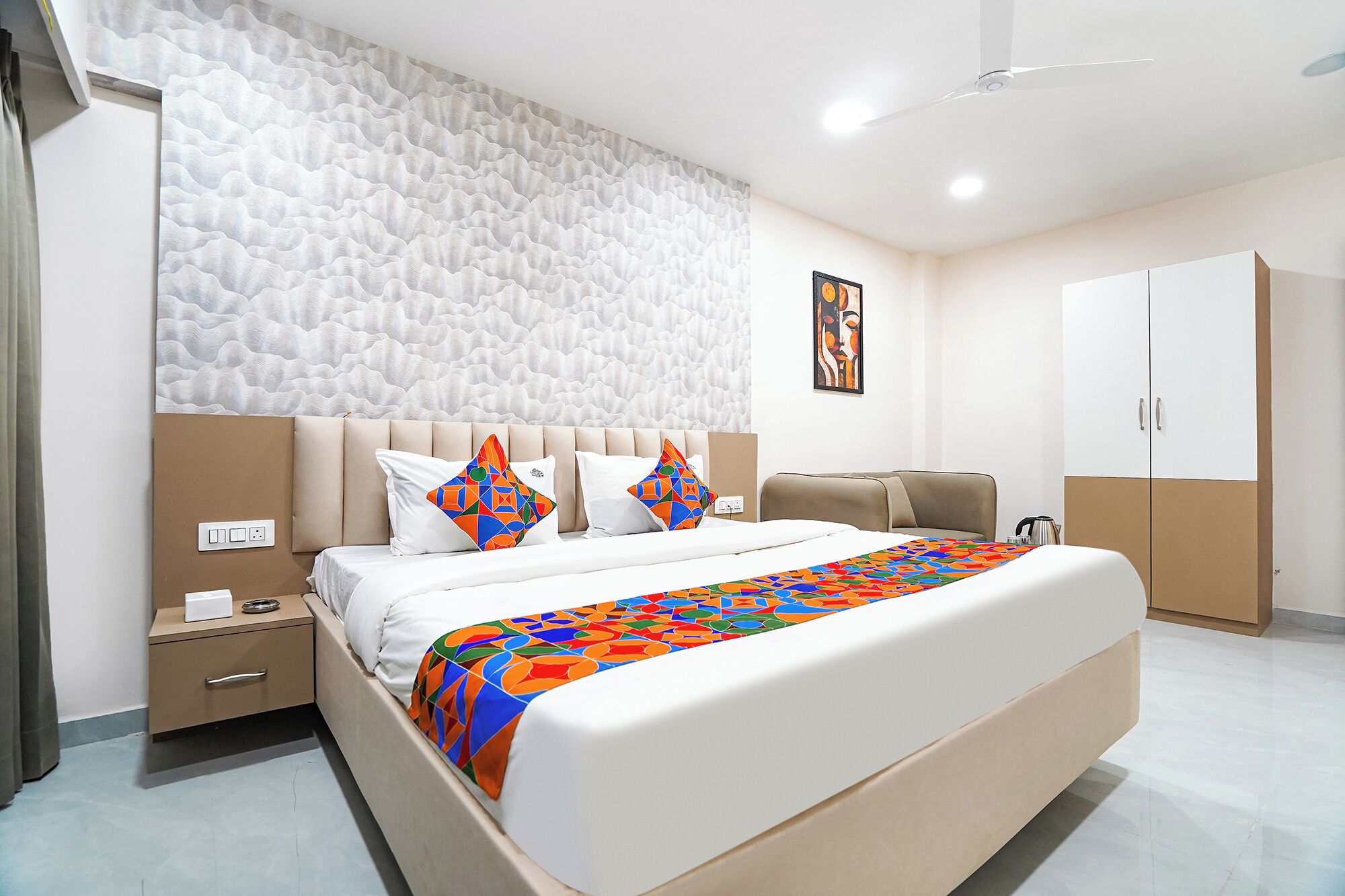 Ein modernes Hotelzimmer mit einem großen Bett, einer Kopfteil mit strukturierter Wand, einem Nachttisch, einem Kleiderschrank und einem Deckenventilator.