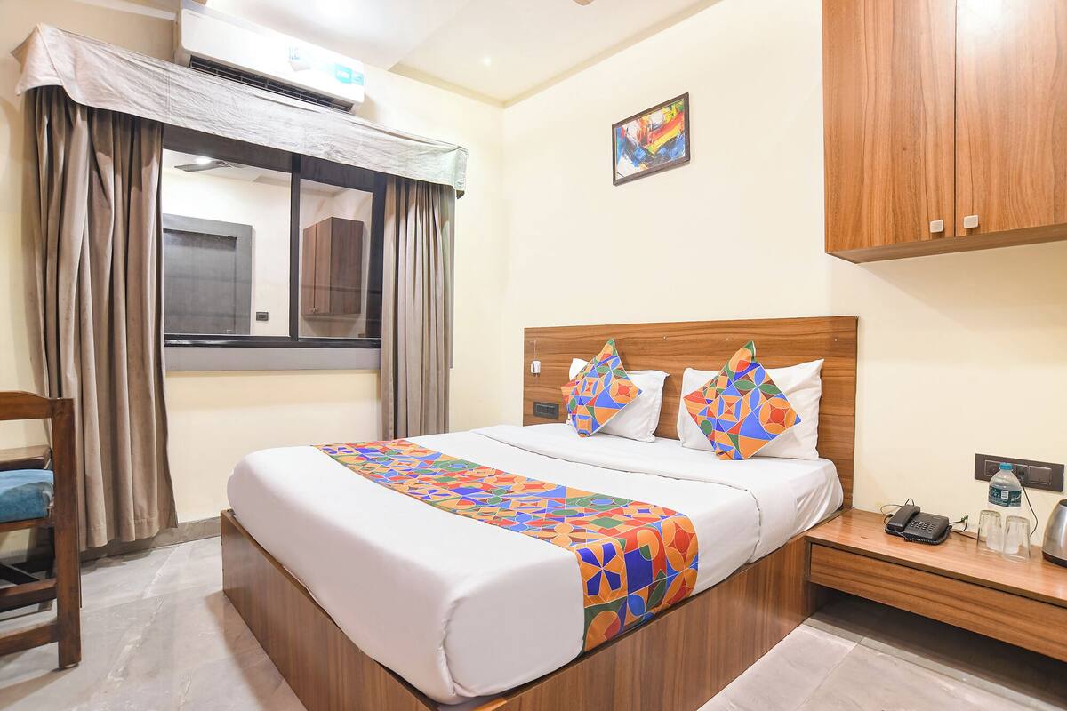 A hotel room with a bed, a wooden headboard, a window with curtains, a bedside table with a phone and glasses, and a wall-mounted air conditioner.