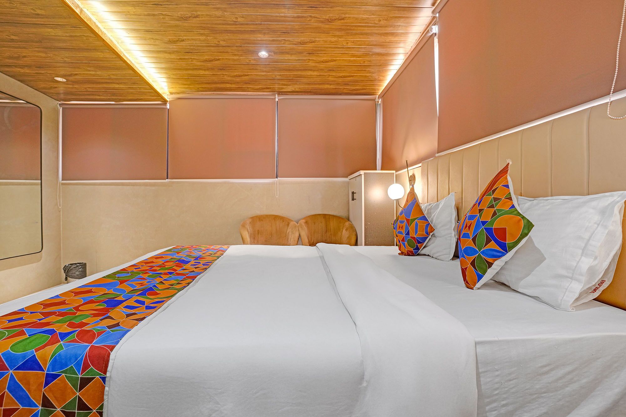 A hotel room with a wooden ceiling, a bed with white linens, two wooden chairs, and a colorful geometric-patterned throw.