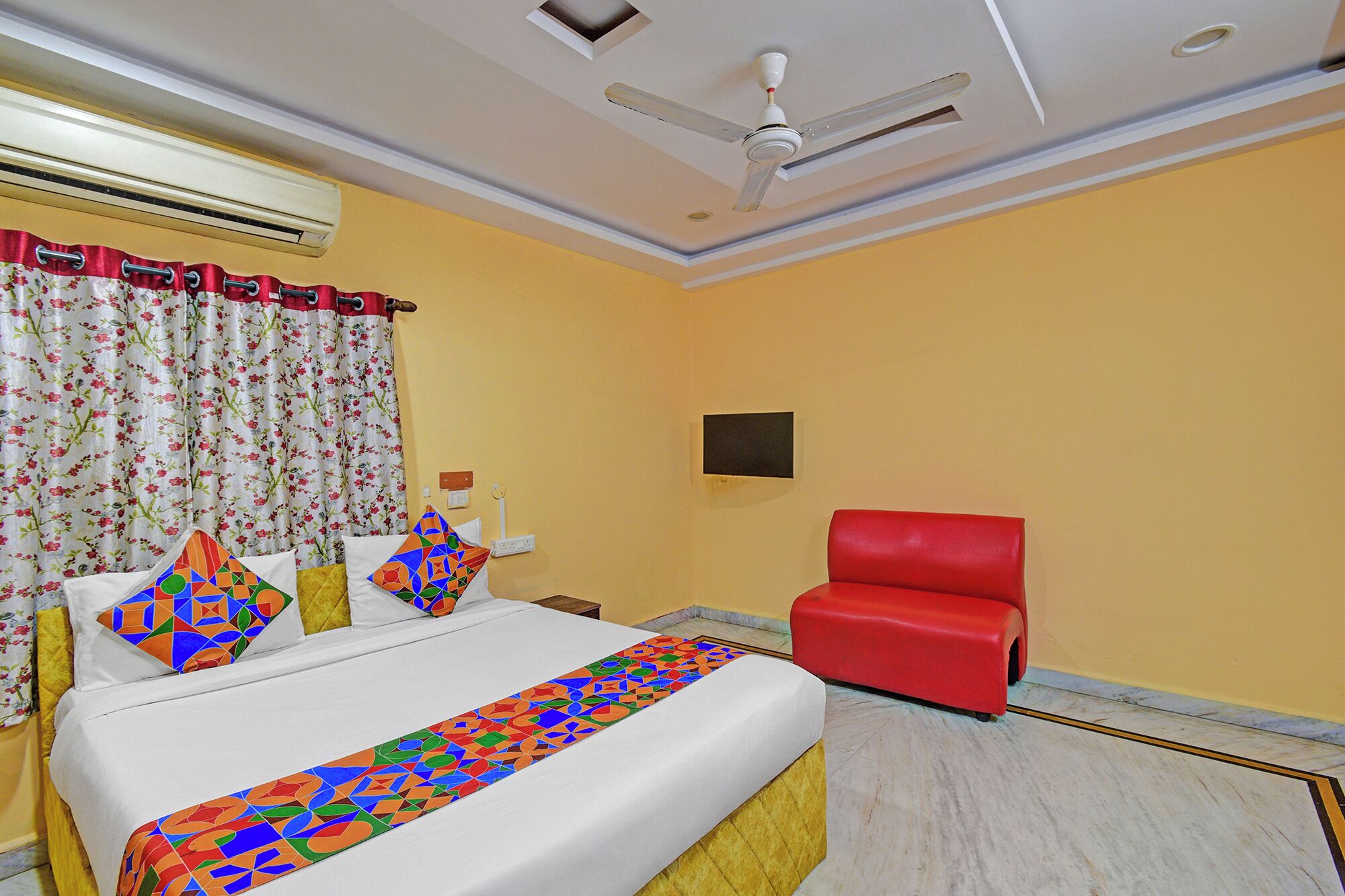 A hotel room with a bed, a red chair, a ceiling fan, and a wall-mounted TV.