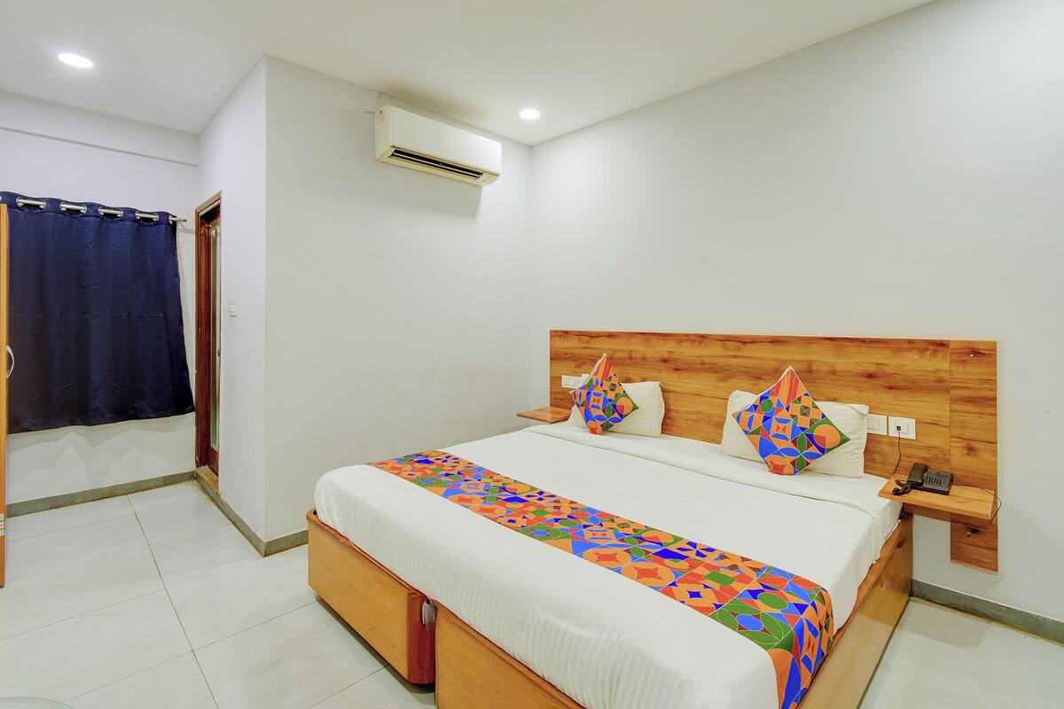 A hotel room with a wooden headboard, a bed with a colorful geometric-patterned bedspread, a bedside table with a phone, and an air conditioning unit on the wall.