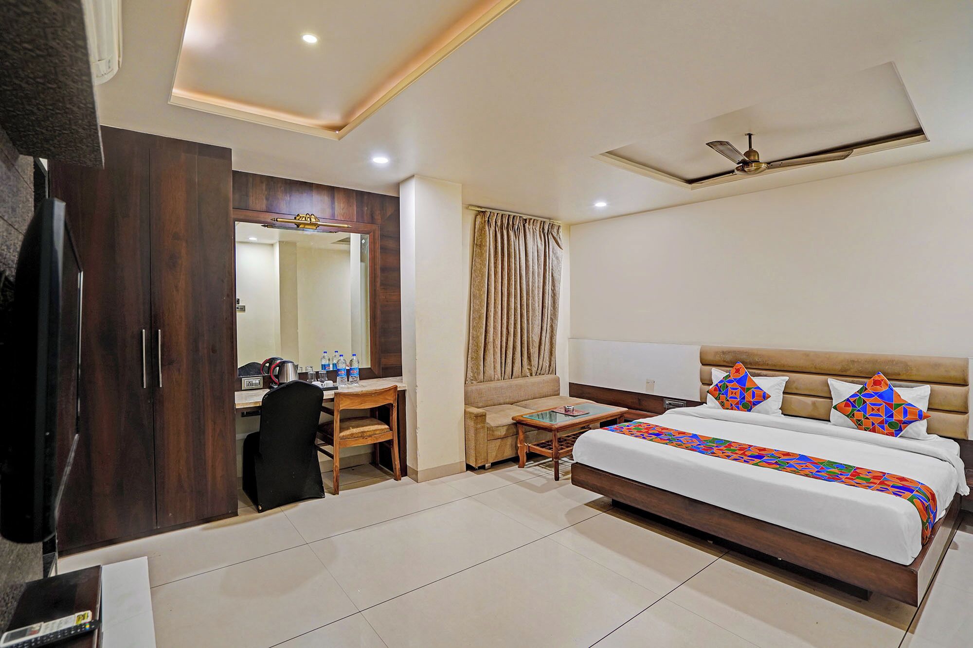 A hotel room with a bed, a desk, a chair, a wardrobe, a mirror, a ceiling fan, and a window with curtains.