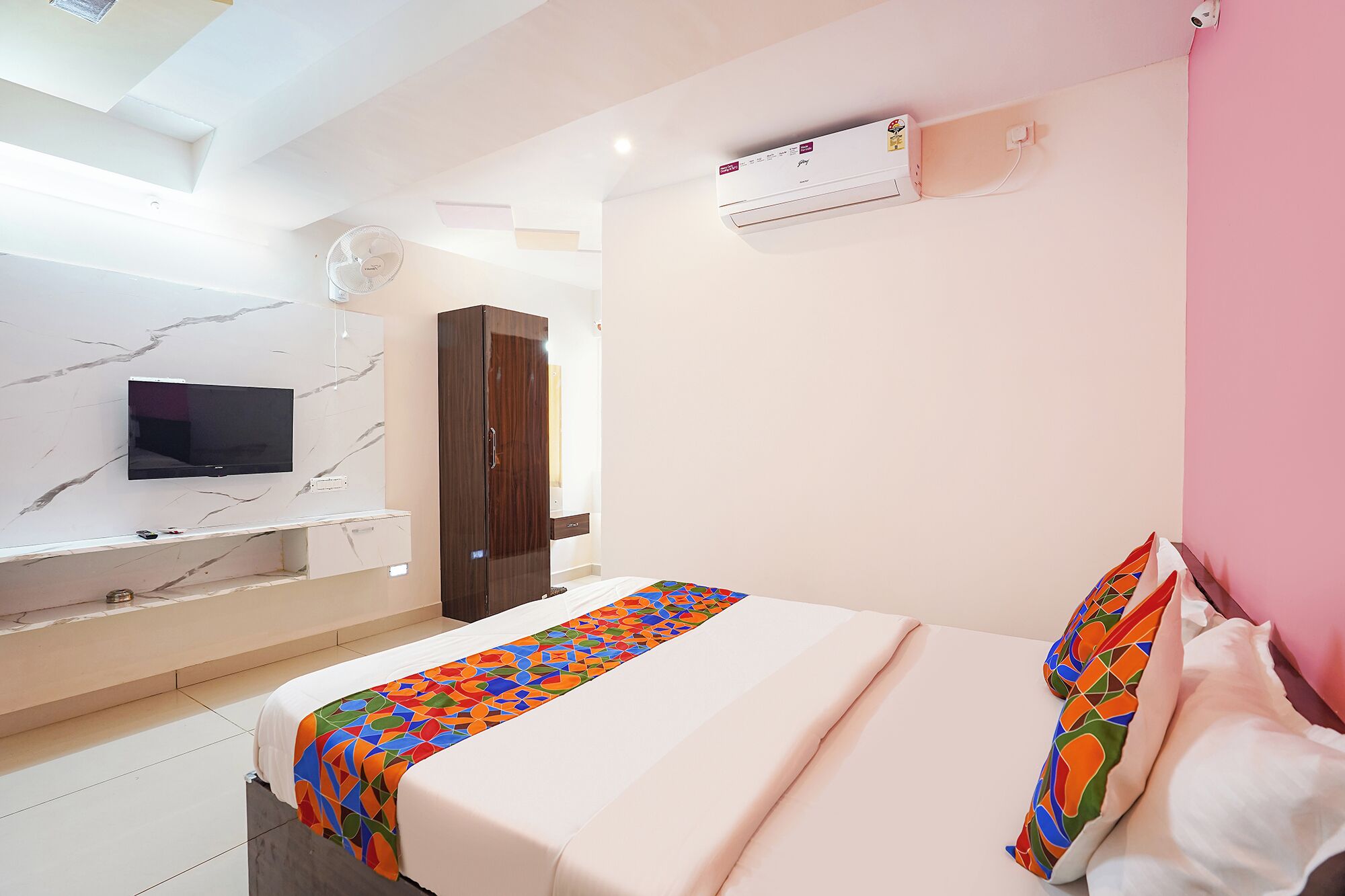 A hotel room with a bed, a TV mounted on the wall, a wardrobe, and an air conditioning unit.