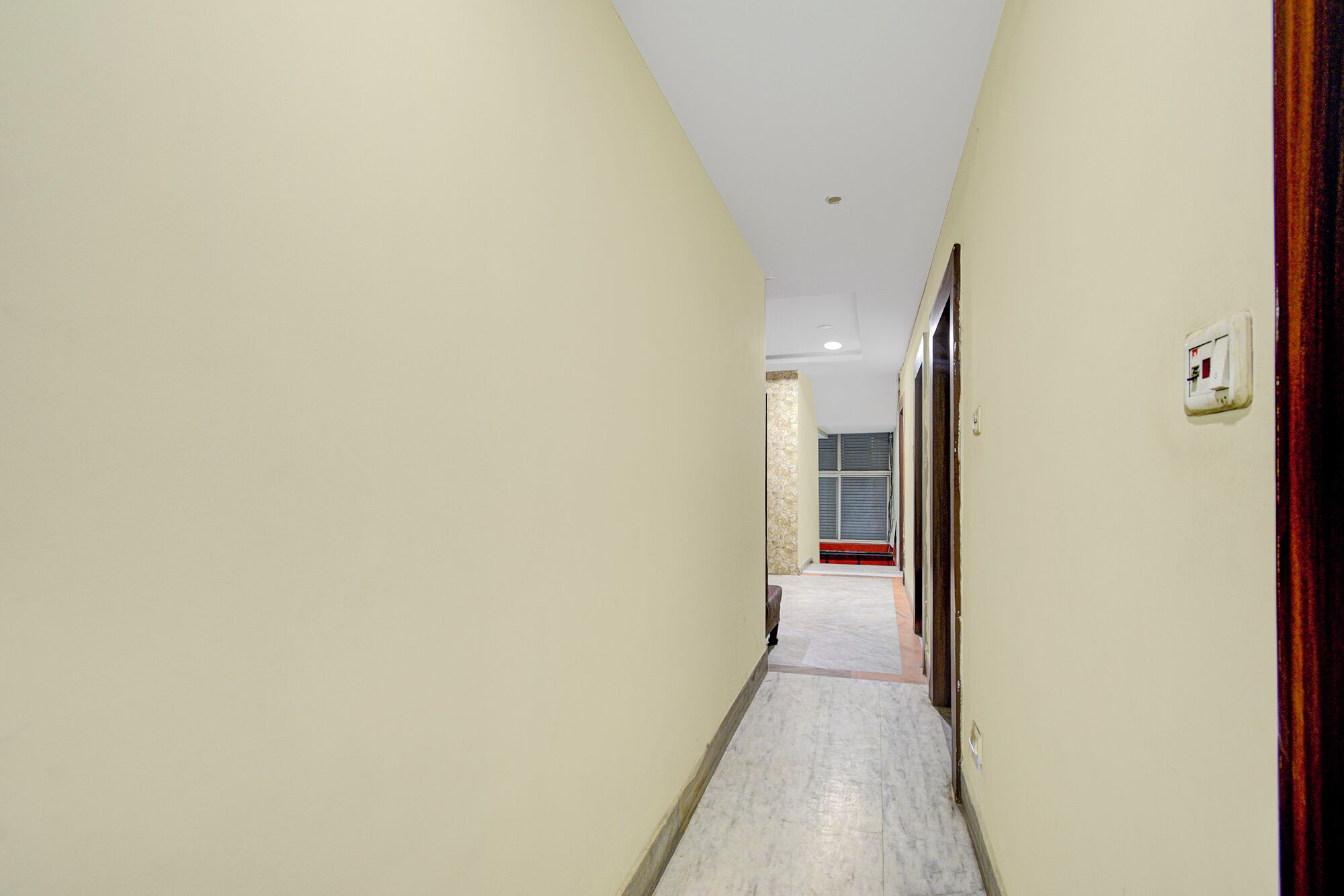 A narrow hallway with light-colored walls and a tiled floor, leading to a room with a window and a door.