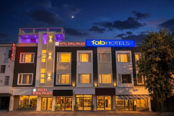 Hotel Suraj Palace com fachada moderna, sinalização iluminada e entrada visível.