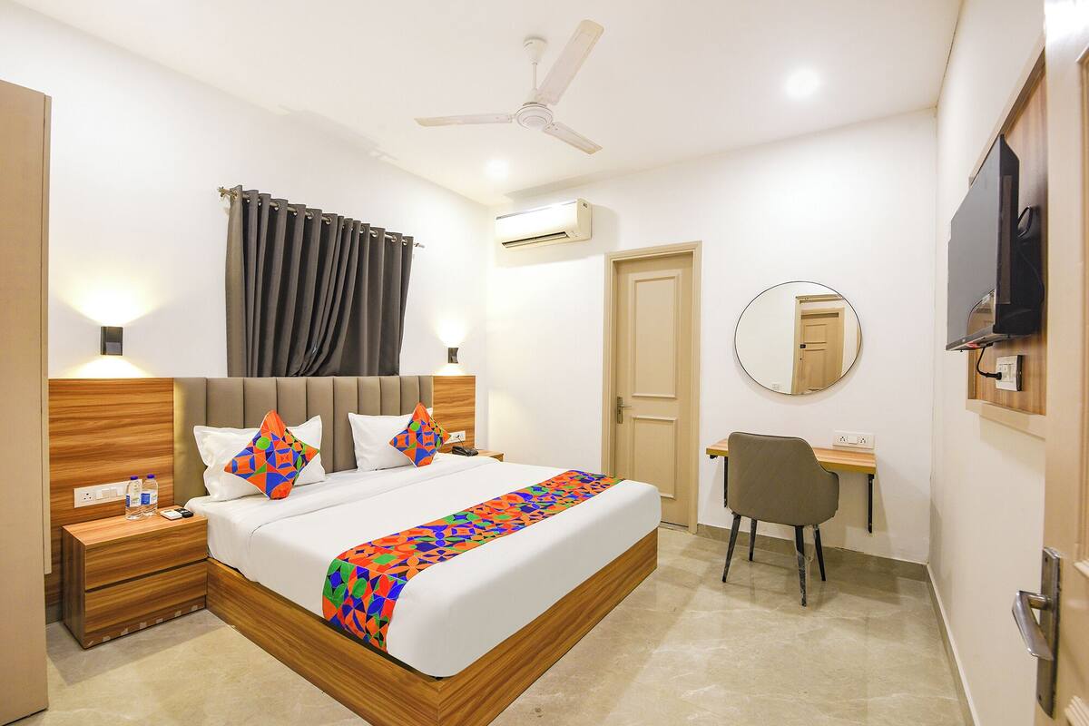 A hotel room with a bed, bedside tables, a desk, a chair, a ceiling fan, an air conditioning unit, a mirror, and a television.