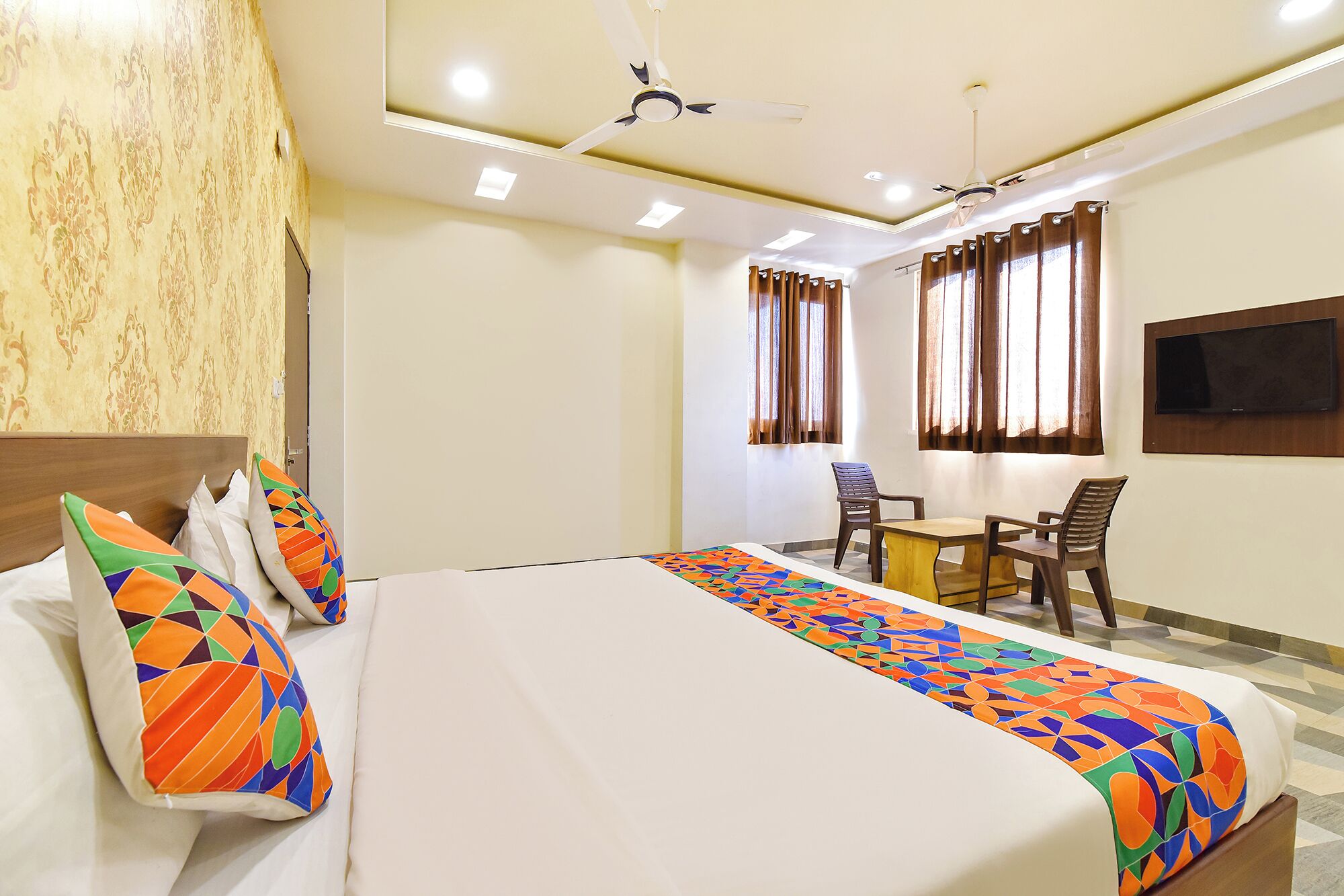 A hotel room with a bed, a desk with two chairs, a ceiling fan, and a TV mounted on the wall.