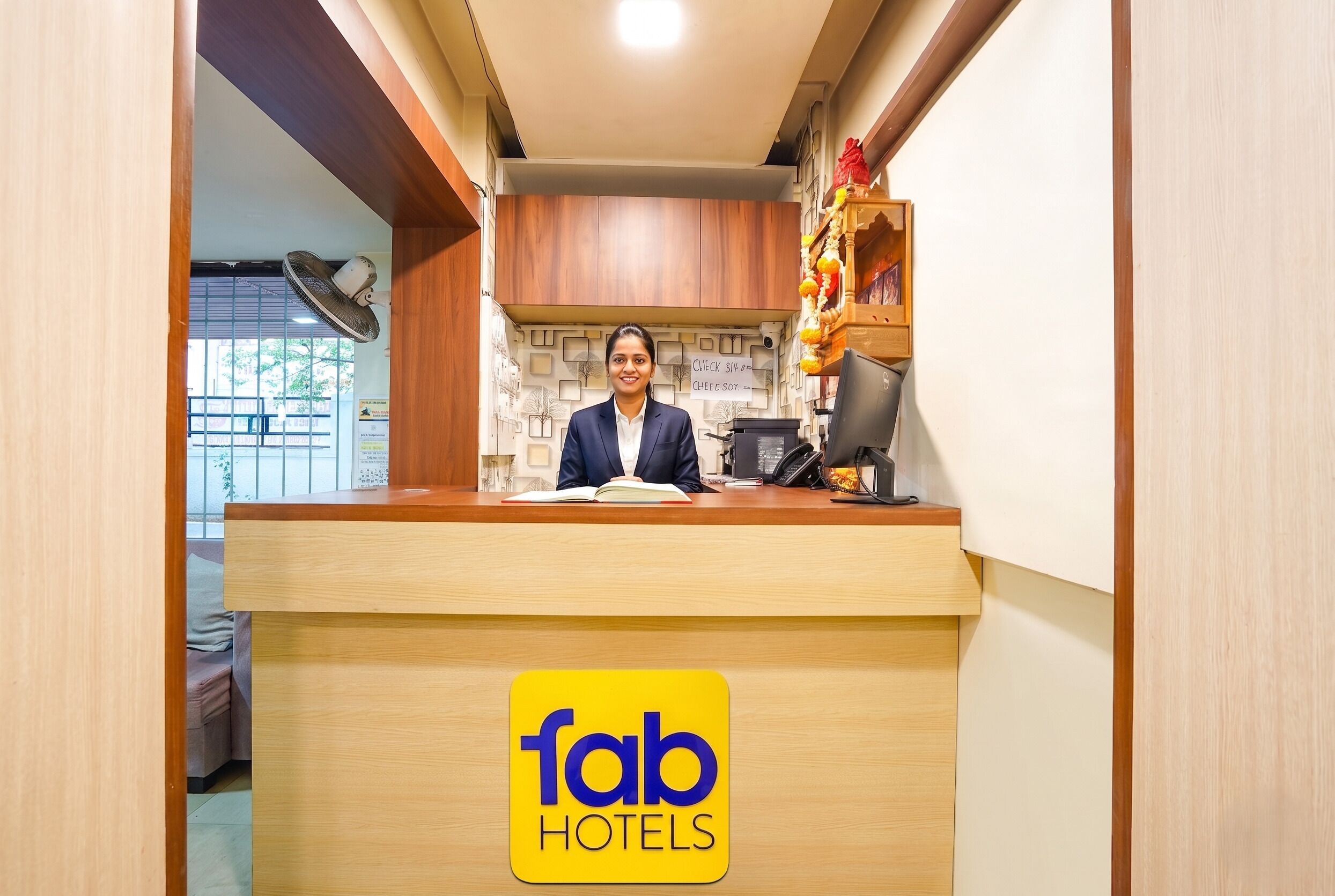 A hotel reception desk with a sign for Fab Hotels, a wooden counter, a fan, and a view of the outside.