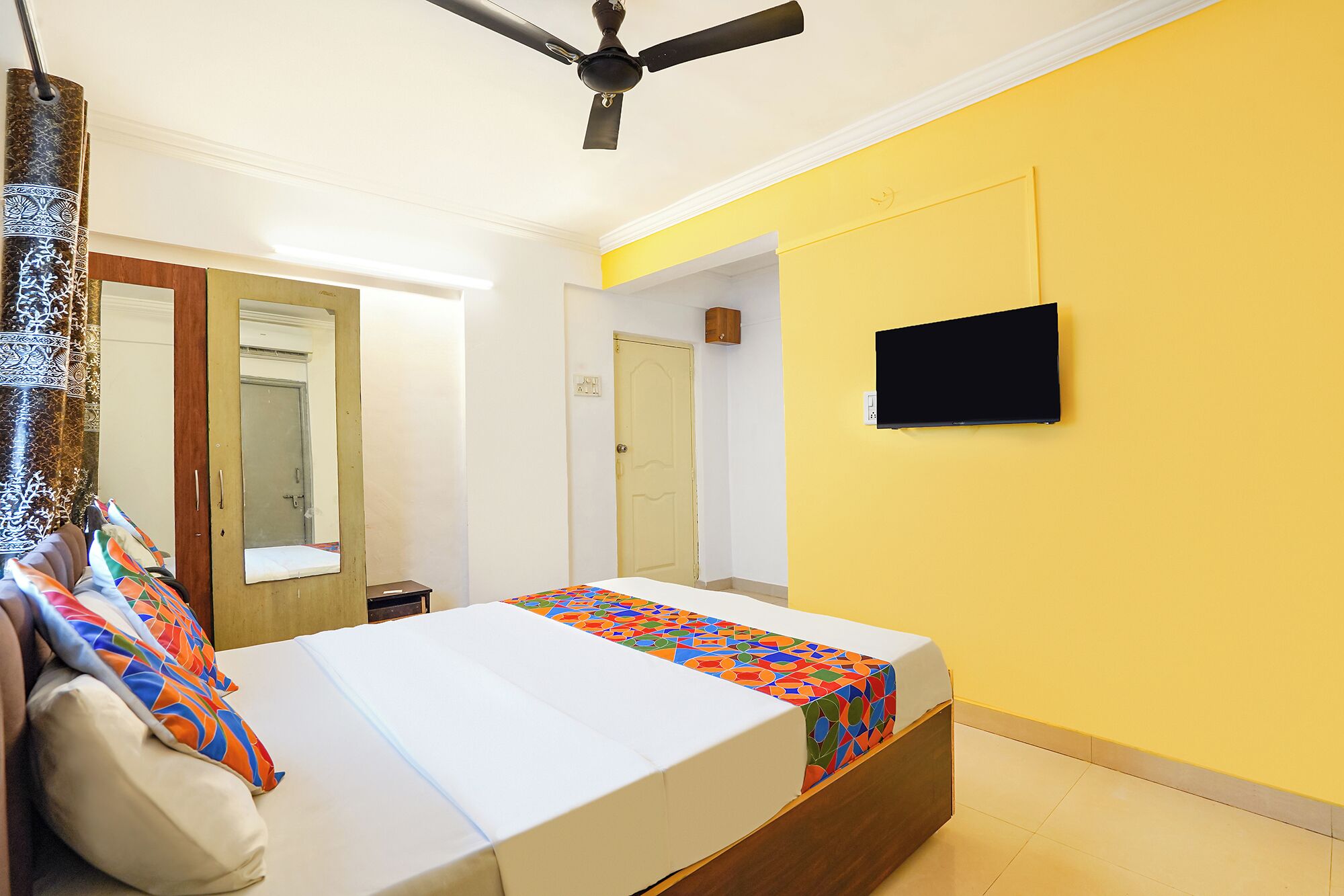 Une chambre à coucher avec un lit, un ventilateur de plafond, un téléviseur fixé au mur et une porte donnant sur une autre pièce.