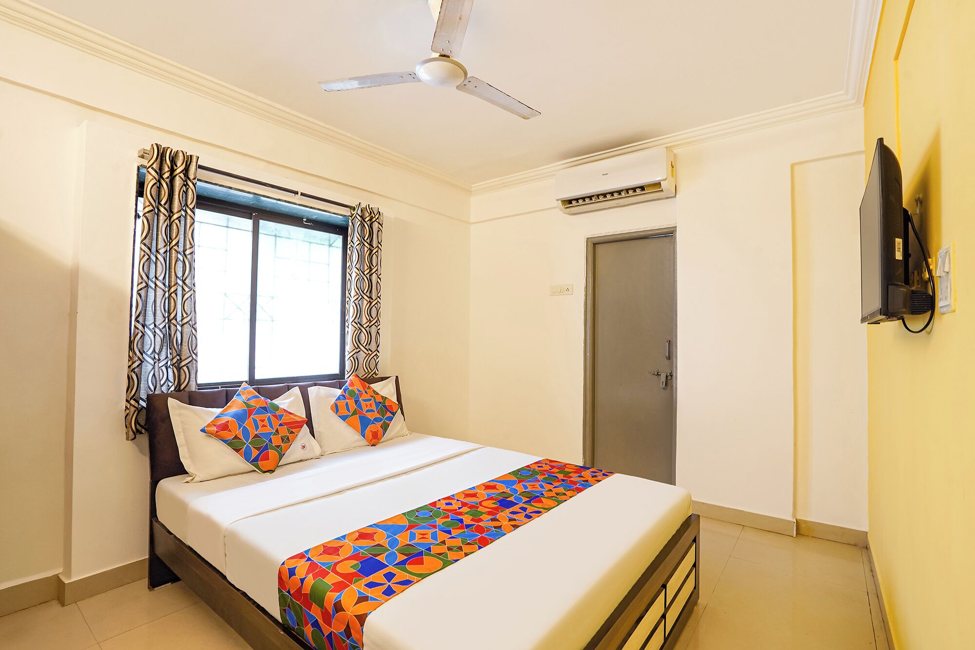 A hotel room with a bed, curtains, a ceiling fan, an air conditioning unit, a television, and a door.