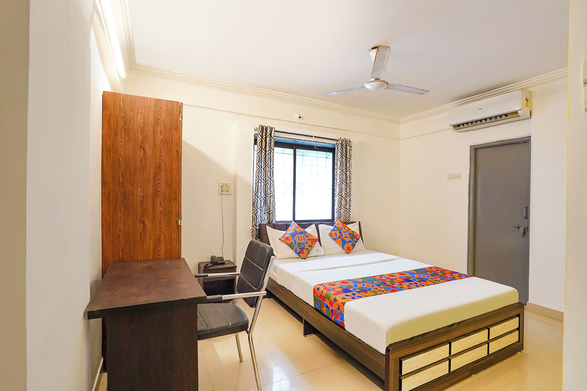 A bedroom with a bed, a desk, a chair, a window with patterned curtains, a ceiling fan, and an air conditioning unit.