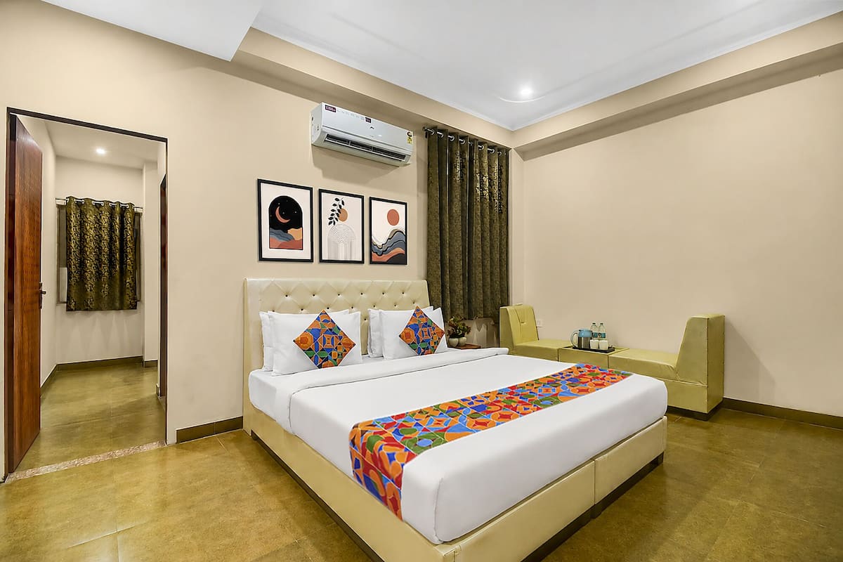 A hotel room with a bed, a bedside table, an air conditioning unit, a mirror, and a window with curtains.