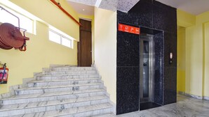 Una scalinata in marmo conduce a un ascensore nero, con un estintore sulla parete e una parete gialla con una porta.