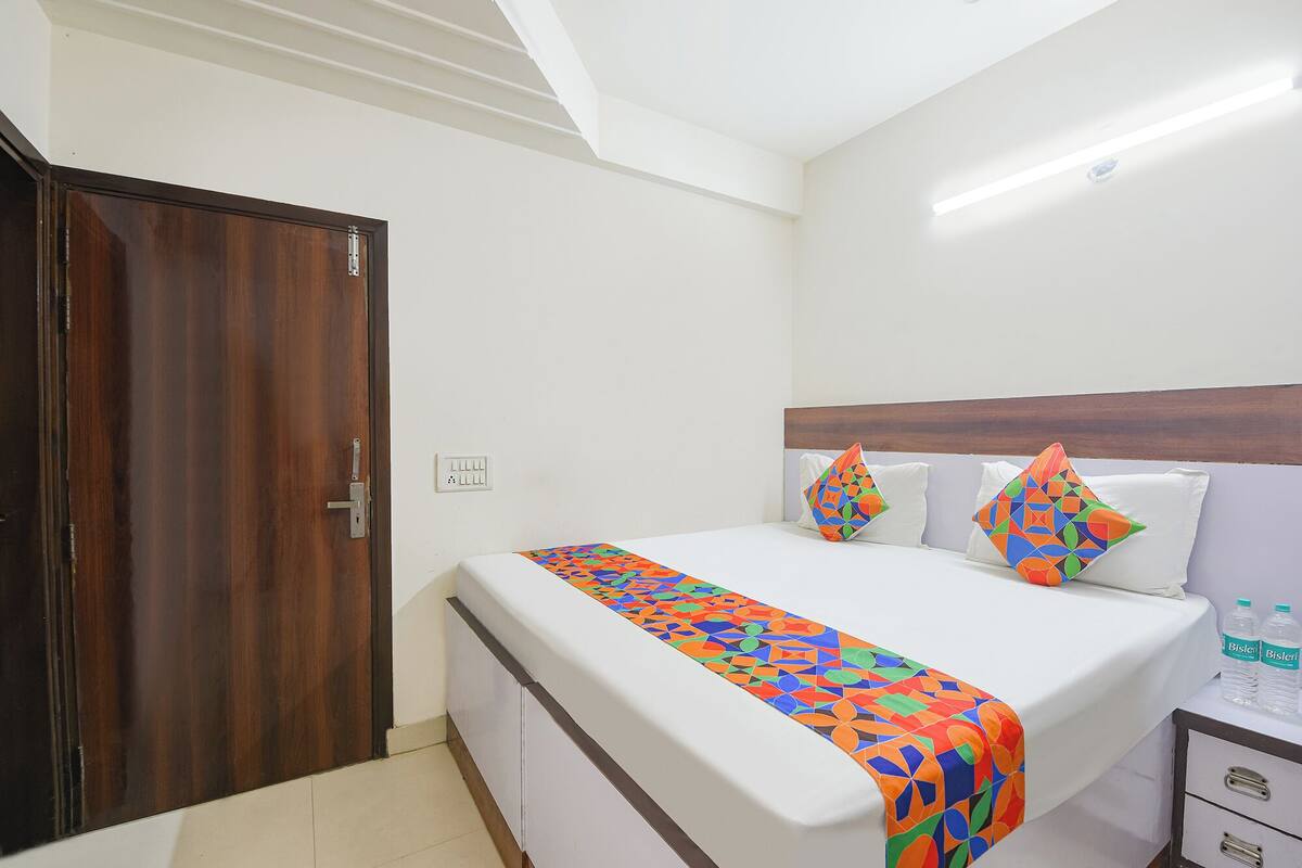 A hotel room with a bed, a wooden door, a white wall, and a nightstand with water bottles.