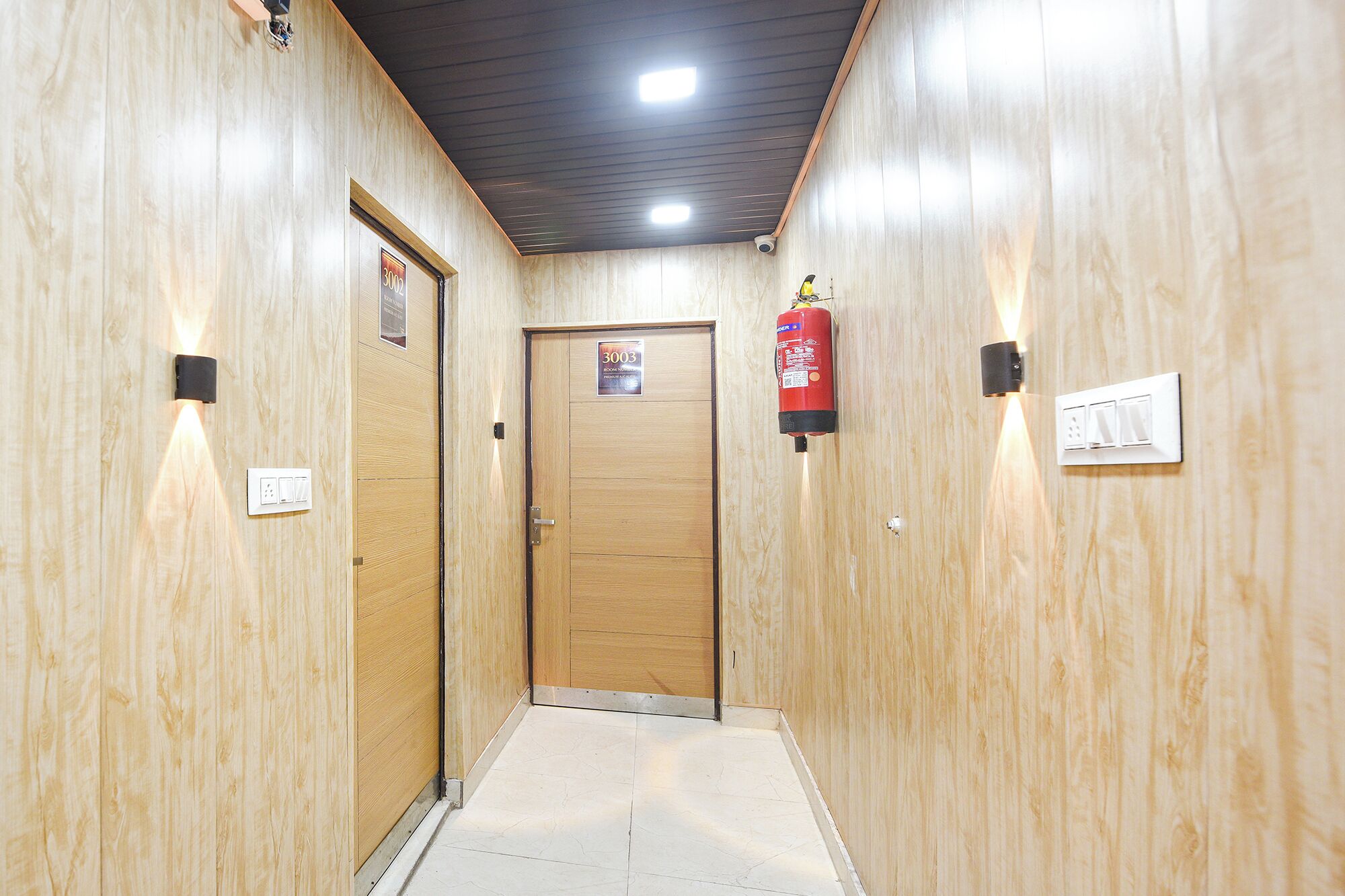A hallway with wooden walls, a door labeled '3003', a fire extinguisher, and a light switch.