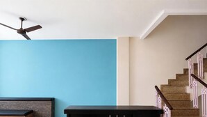 A room with a blue wall, a ceiling fan, a staircase with pink railings, and a bench. - Via A Square Residency (New Delhi)