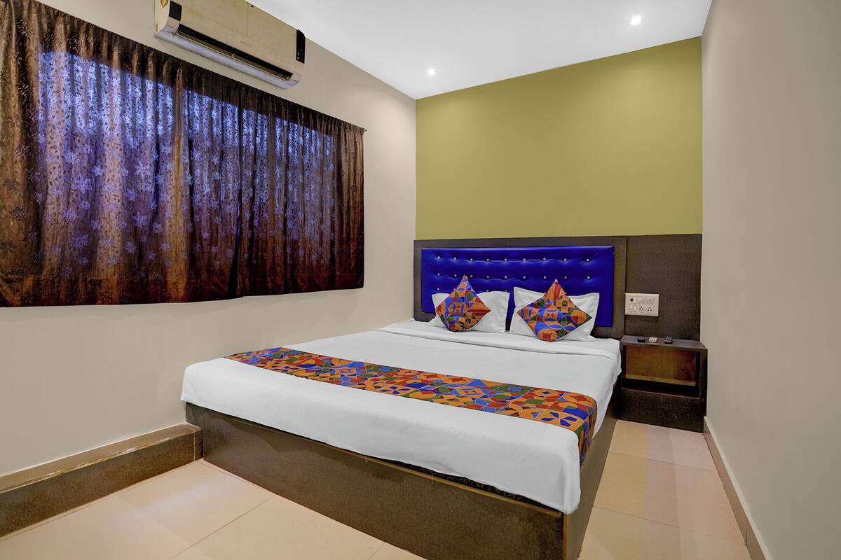 A hotel room with a bed, a nightstand, an air conditioning unit, and a window with curtains.