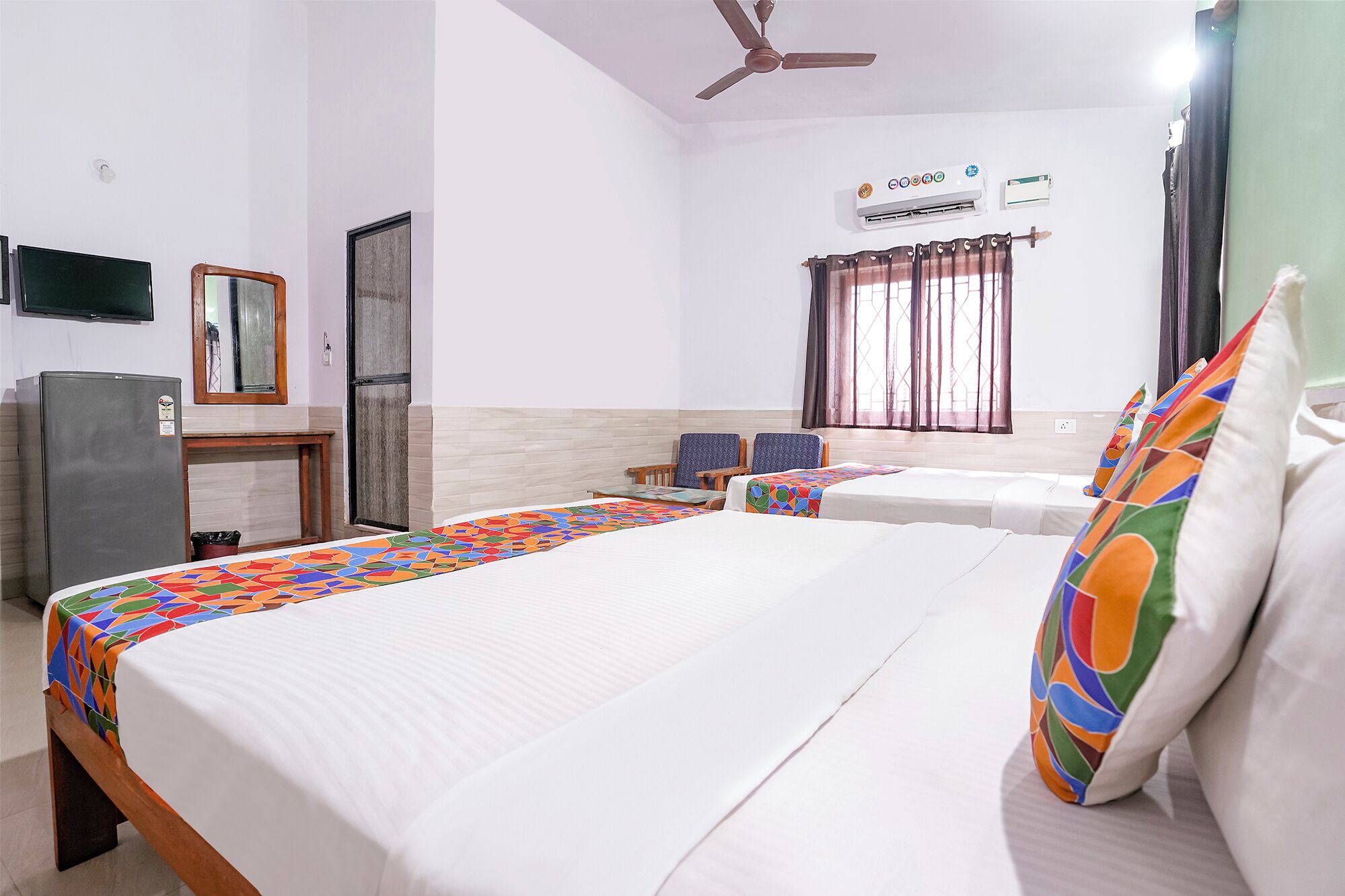 Quarto de hotel com cama, duas cadeiras, escrivaninha, geladeira, ar-condicionado e ventilador de teto.