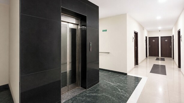A hallway with a black elevator, a white wall with an exit sign, and a door with a number. - Via Cosy Stay (New Delhi)