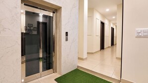 A modern elevator with a digital display, a clean hallway with a green artificial grass area, and a wall-mounted electrical outlet. - Via Ak Residency (New Delhi)