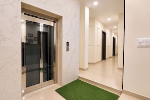 A modern elevator with a digital display, a clean hallway with a green artificial grass area, and a wall-mounted electrical outlet. - Via Ak Residency (New Delhi)