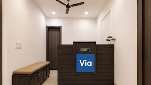 A reception area with a dark wood reception desk, a blue sign with 'Via' on it, a bench, and a ceiling fan. - Via Ak Residency (New Delhi)