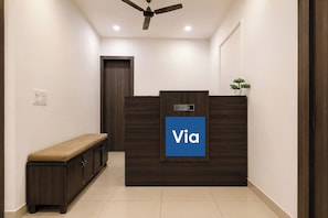 A reception area with a dark wood reception desk, a blue sign with 'Via' on it, a bench, and a ceiling fan. - Via Ak Residency (New Delhi)