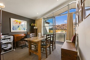 A dining area with a wooden table and chairs, a desk with a printer, a framed painting, and a view of mountains through large windows. - Stylish Sunny Apartment with Mt Roy Views, Balcony & Coffee Machine | Walk to Breweries, Town & Lake (Wānaka)