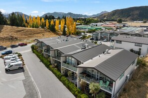 A modern building with a parking lot, surrounded by trees and mountains. - Stylish Sunny Apartment with Mt Roy Views, Balcony & Coffee Machine | Walk to Breweries, Town & Lake (Wānaka)