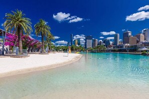 A beach with clear water, palm trees, and a city skyline in the background. - LVL72 Breathtaking Views with Parking, Pool & Gym (Brisbane City)