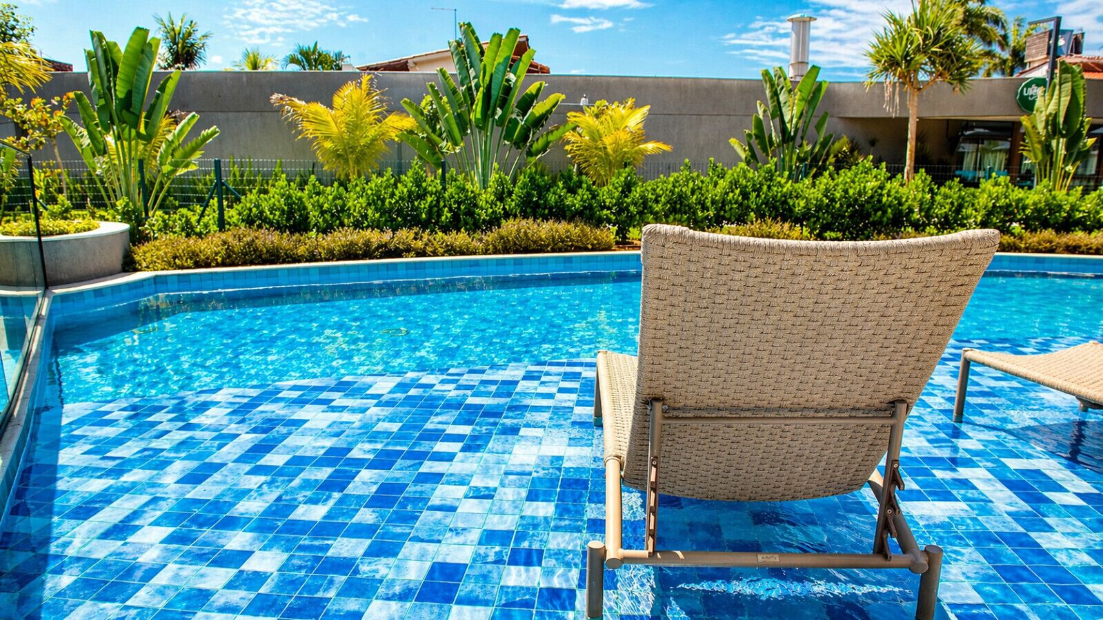 Una piscina con baldosas azules a cuadros, rodeada de exuberante vegetación y una tumbona.