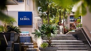 A hotel entrance with a sign reading 'THE SEBEL,' a staircase, and a planter with a palm tree.