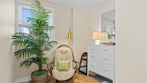 Una habitación con una silla de mimbre, una planta en maceta, una lámpara y un tocador blanco.