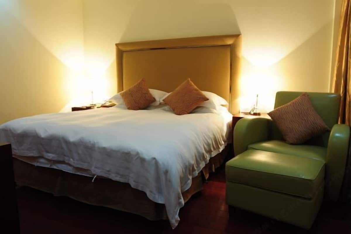 Habitación de hotel con una cama, dos almohadas, una mesita de noche, un sillón verde y una lámpara.