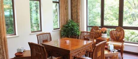 A dining area with a wooden table and chairs, large windows with a view of greenery, and a hanging light fixture.