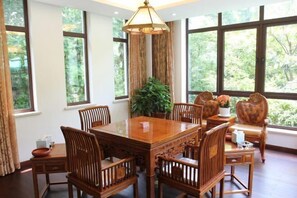 Un comedor con mesa y sillas de madera, ventanas amplias con vistas a la vegetación y una lámpara colgante.