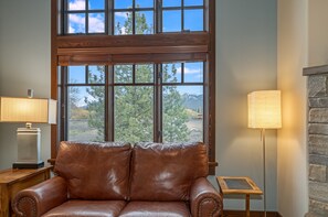 A room with a brown leather sofa, a lamp, and a view through large windows. - New! Cozy Craftsman Condo | Walk to Downtown Whitefish, Gas Fireplace & Mountain Views (Whitefish)